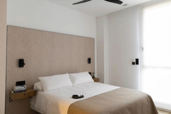 Cama doble con cabecero beige y pared blanca en Alma Demetrio.