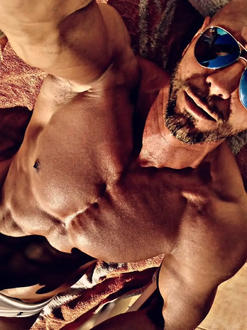 Hombre musculoso tomando un selfie al sol con gafas de sol y torso definido.