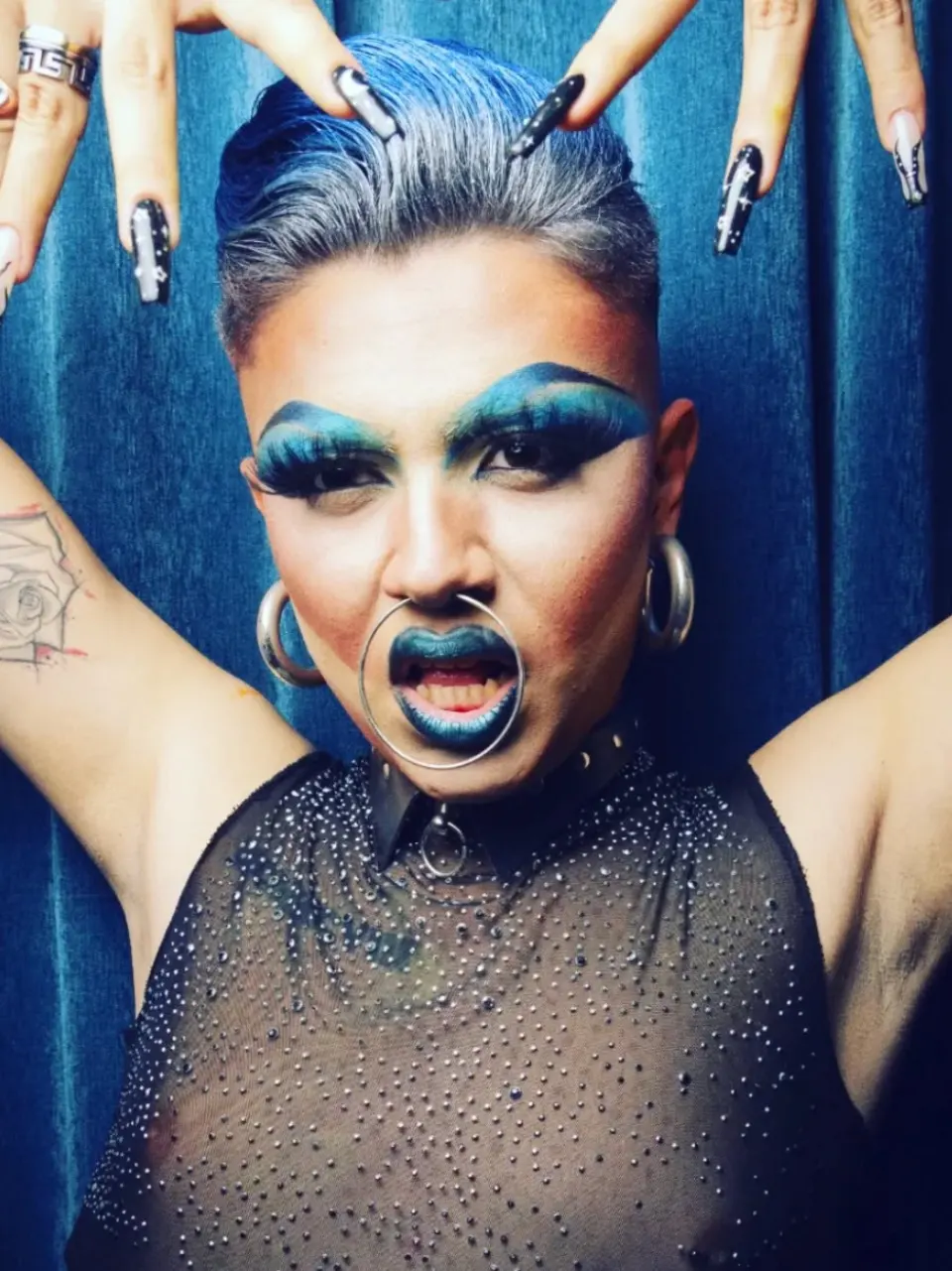 Drag queen conceptual con maquillaje azul, expresión intensa y estilismo alternativo posando frente a un fondo azul.