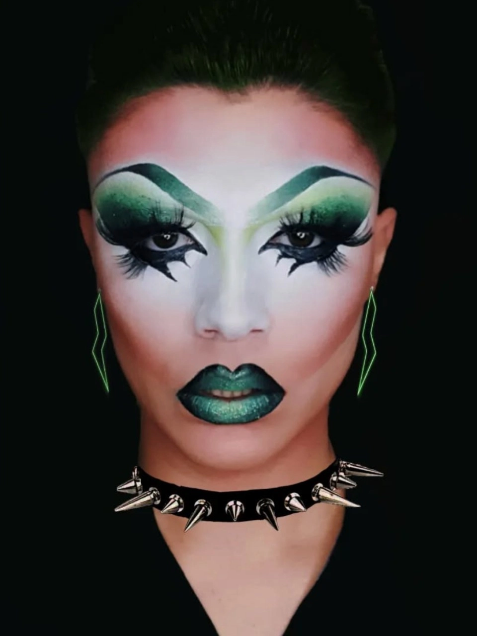 Drag queen conceptual con maquillaje verde artístico, labios metalizados y collar de pinchos sobre fondo negro.
